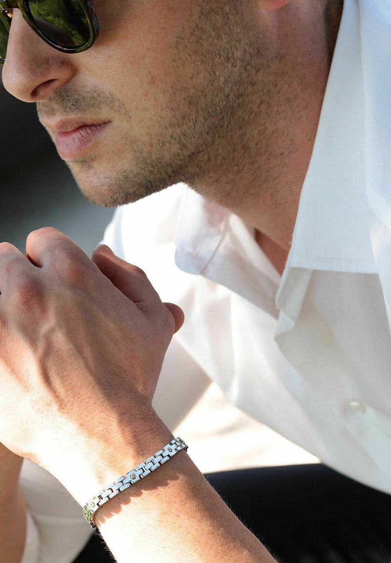 Bracciale in metallo argentato con design a catena, superfici lucidate e accenti circolari. Indossato su un polso, sopra una camicia di colore chiaro.