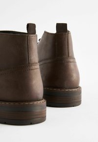 Botas de cuero marrón hasta el tobillo que presentan un acabado texturizado, pestañas reforzadas y una suela de madera en contraste con detalles de costura.