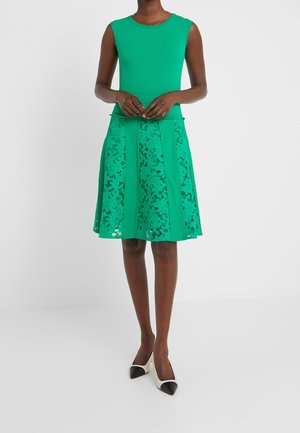 Zielona sukienka bez rękawów z fakturowaną górą i panelami spódnicy z kwiatowej koronki, noszona z czarno-białymi czółenkami na szpic na modelce stojącej.
