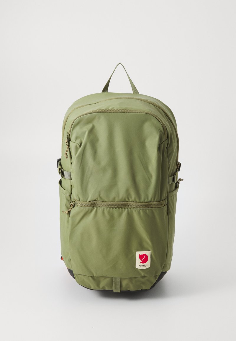 Mochila verde de nylon com dois bolsos frontais com fecho, alças laterais ajustáveis e um emblema vermelho na parte inferior. Apresenta uma pega na parte de cima.