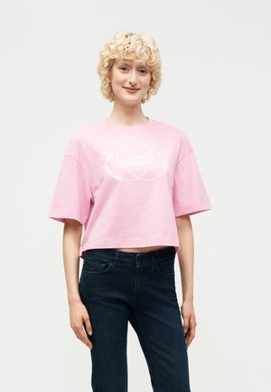 Fiatal személy rövid, göndör szőke hajjal, laza, rózsaszín, rövidített pólót visel fehér nyomattal és sötétkék farmernadrágot, egy egyszerű háttér előtt áll.