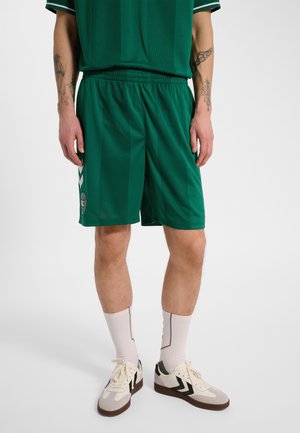 Person iført grønne sportsshorts og top, hvide sokker, beige og sorte sneakers, står med hænderne afslappet langs siderne.