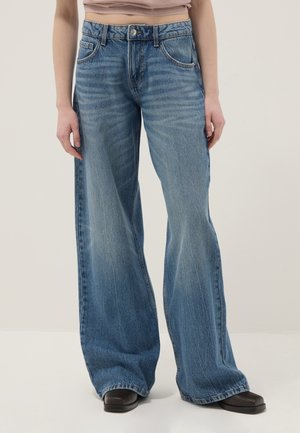 Person trägt hoch taillierte, weit geschnittene blaue Jeans und schwarze Schuhe mit quadratischer Spitze, steht vor einem schlichten hellen Hintergrund.