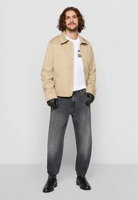 Giacca beige in cotone con design colletto, indossata sopra una t-shirt bianca, abbinata a jeans grigi e stivali in pelle nera.