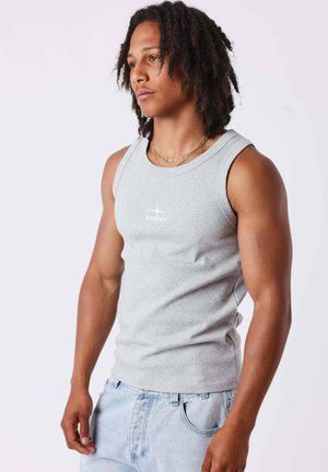 Giovane uomo con dreadlocks che indossa un top grigio aderente senza maniche e jeans azzurri, in piedi contro uno sfondo bianco uniforme.