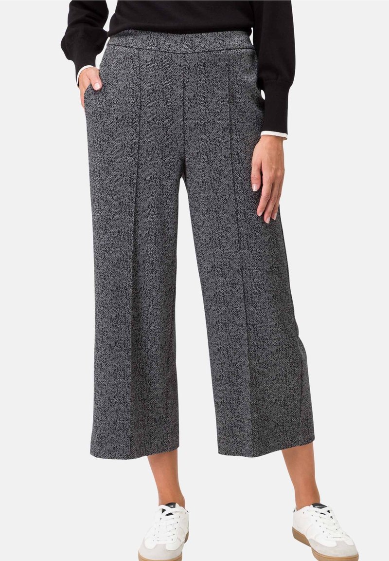 Femme portant un pantalon gris à jambes larges et courts avec un motif subtil, un haut noir à manches longues et des baskets blanches, debout avec une main dans la poche.