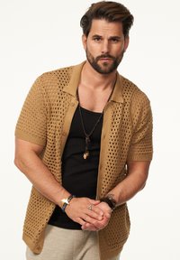 Beige Strickjacke mit kurzen Ärmeln und offenen Maschen, kombiniert mit einem schwarzen Tanktop. Die Accessoires umfassen eine geschichtete Halskette und Armbänder.