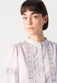 Gaiši rozā blūze ar augsto apkakli, kurai ir sarežģīta balta mežģīņu apdare un pogu aizdare priekšā. Puff piedurknes akcentē dizainu.