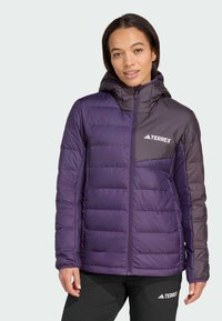 Eine Frau trägt eine zweifarbige lila Terrex gepolsterte Jacke mit Kapuze und schwarze Terrex-Hosen. Sie steht vor einem einfarbigen hellen Hintergrund.