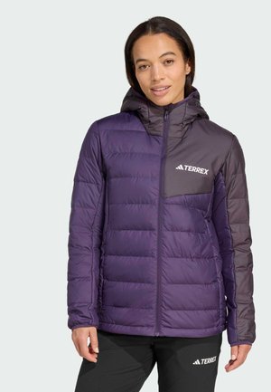 Woman wearing a two-tone purple Terrex padded jacket with hood and black Terrex pants, standing against a plain light background.