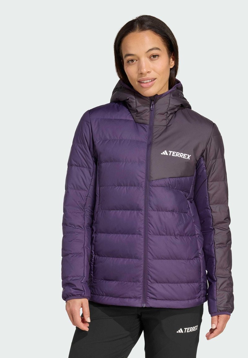 Eine Frau trägt eine zweifarbige lila Terrex gepolsterte Jacke mit Kapuze und schwarze Terrex-Hosen. Sie steht vor einem einfarbigen hellen Hintergrund.