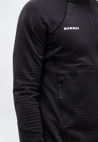 Κοντινό πλάνο ενός ατόμου που φοράει μαύρο μπουφάν Mammut με φερμουάρ, με ορατή τσέπη με φερμουάρ στο πλάι.