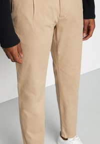Beige trousers with a tailored fit, featuring a smooth texture and subtle pleats. Paired with white sneakers, contouring the ankles.