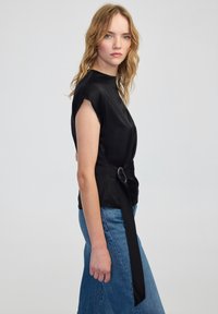 Giovane donna con capelli biondi ondulati che indossa una maglietta senza maniche nera con una cintura e jeans blu, in piedi contro uno sfondo semplice.