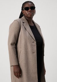 Manteau en mélange de laine beige clair avec un col cranté, une fermeture à double bouton, des poches latérales et une texture lisse. Porté sur un haut noir.