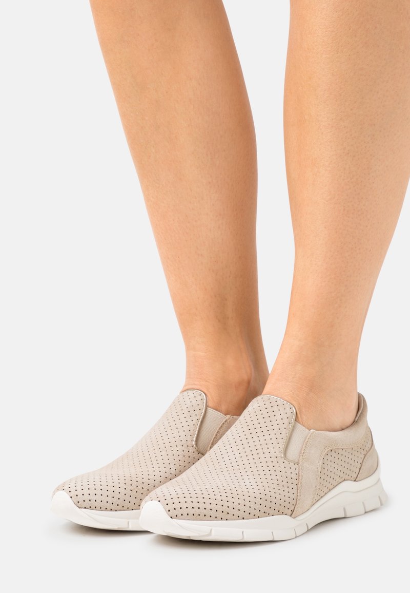 Chaussures décontractées beige à enfiler avec un design perforé portées pieds nus sur fond blanc.