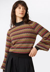 Mehrfarbiger strukturierter Pullover mit Zickzackmuster, langen Ärmeln und einem schwarzen gerippten Kragen sowie Saum. Kombiniert mit schwarzen Hosen.