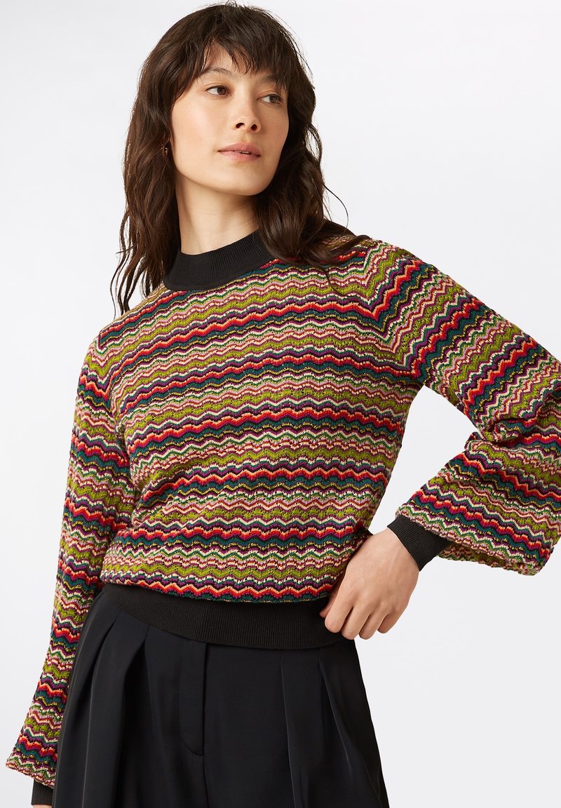 Mehrfarbiger strukturierter Pullover mit Zickzackmuster, langen Ärmeln und einem schwarzen gerippten Kragen sowie Saum. Kombiniert mit schwarzen Hosen.
