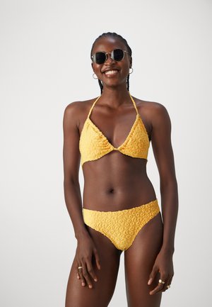 Kollane bikiin, millel on tekstureeritud ja mustriline kangas, halter kaelusega tops ja madala vöökoha aluspüksid. Lisadeks ringikujulised prillid.