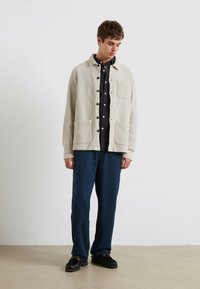 Giovane in piedi all'interno che indossa una giacca beige con bottoni, una camicia nera, pantaloni blu e scarpe nere, mentre guarda in basso.