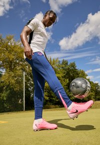 Kék sportnadrág rózsaszín részletekkel, fehér, rövid ujjú póló és rózsaszín focicipő, amint egy fekete és szürke Nike focilabdát irányít.