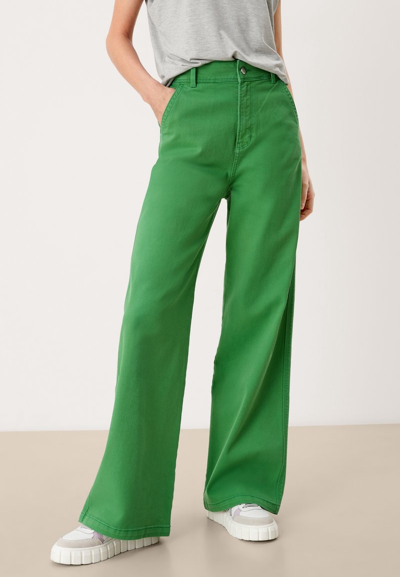 Personne portant un pantalon large vert vif, un t-shirt gris rentré et des baskets blanches, debout les mains dans les poches devant un fond uni.
