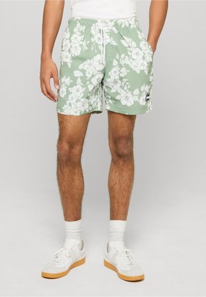 Bărbat purtând pantaloni scurți verzi cu imprimeu floral alb, șosete albe și adidași albi, stând în fața unui fundal simplu gri deschis.
