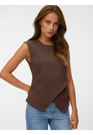 Femme portant un haut marron sans manches avec un ourlet asymétrique associé à un jean bleu clair, posant avec une main sur la hanche.