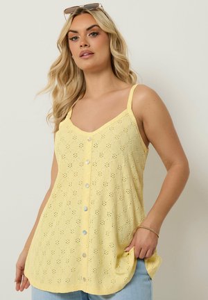 Blond vrouw met zonnebril op het hoofd, mouwloos geel topje met knopen en oogjespatroon, lichtblauwe spijkerbroek en gouden armband om de rechterpols.