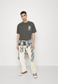 Gray cotton t-shirt with turquoise graphic, cream-colored pants, and a light blue plaid shirt tied around the waist. Brown sneakers complete the look.