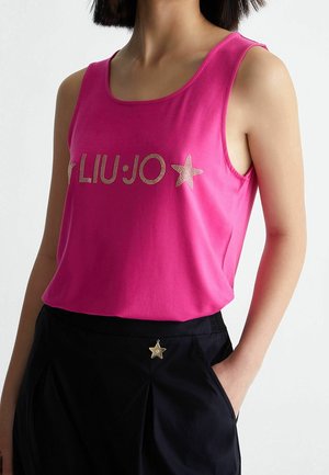 Vrouw draagt een roze mouwloos topje met gouden sterrenstuds en een zwarte broek met een gouden sterrenknop op de tailleband, hand in de zak.