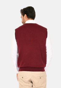 Gilet lavorato a maglia color bordeaux con texture a coste e senza maniche, indossato sopra una camicia bianca con colletto, presentando un colore uniforme senza motivi.