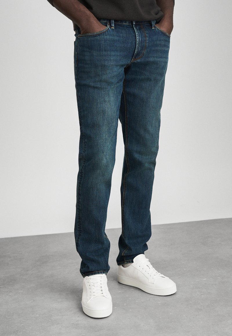 Sötétkék, slim fit farmernadrágot és fehér sportcipőt viselő személy áll egy szürke padlón, a kezét a zsebében tartva.