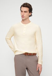 Ανοιχτό κίτρινο μακρυμάνικο πουκάμισο τύπου henley με πέντε κουμπιά, συνδυασμένο με γκρι παντελόνι και καφέ ζώνη. Απλός, εφαρμοστός σχεδιασμός και απαλό ύφασμα.