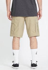 Pantalones cortos beige de estilo cargo con dos bolsillos laterales, ajuste relajado y detalles de costura visibles. Llevados con zapatillas negras y calcetines blancos hasta el tobillo.