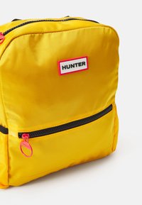Geltonas nailonas kuprinė su didele priekinės kišenės, juodu užtrauktuku ir rožiniu užtrauktuko traukikliu. Yra balta etiketė su žodžiu "HUNTER" juodu tekstu.