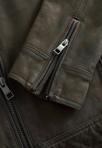 Braune Lederjacke mit Detailansicht auf den Reißverschluss und die Nähte. Das Material hat eine strukturierte Oberfläche mit subtilen Abnutzungsspuren und einer klaren Form.