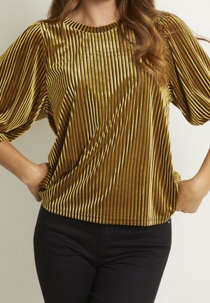 Femme portant un haut en velours à rayures verticales dorées et noires avec des manches bouffantes, associée à un pantalon noir, les mains sur les hanches.