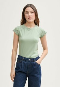 Gaiši zaļš pieguļošs tops ar īsām frillēm uz piedurknēm, savienots ar augsto jostasvietu tumši ziliem džinsiem, kas izceļas ar uzšuvēm un kabatām.