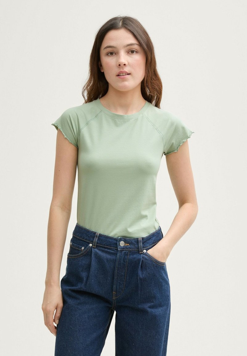 Gaiši zaļš pieguļošs tops ar īsām frillēm uz piedurknēm, savienots ar augsto jostasvietu tumši ziliem džinsiem, kas izceļas ar uzšuvēm un kabatām.