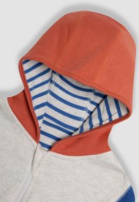 Hoodie mit einer orangen Kapuze, weißem Körper und blauen Akzenten. Innen umfasst er einen blau-weiß gestreiften Stoff. Weicher Baumwolltextil. Mit Reißverschluss.