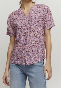 Lilla blomster kortærmet bluse med V-hals, der har flerfarvede blomstermønstre, en knapdetalje og en buet kant.