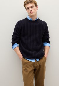 Marineblauer Strickpullover mit Rippenstruktur, Rundhalsausschnitt und aufgerollten Ärmeln über einem hellblauen Hemd. Beige Hose vervollständigt das Outfit.