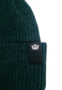 Berretto in maglia verde con texture a coste, caratterizzato da una toppa in tessuto nero con un logo chiaro, che mostra un design senza soluzione di continuità e uno stile funzionale.