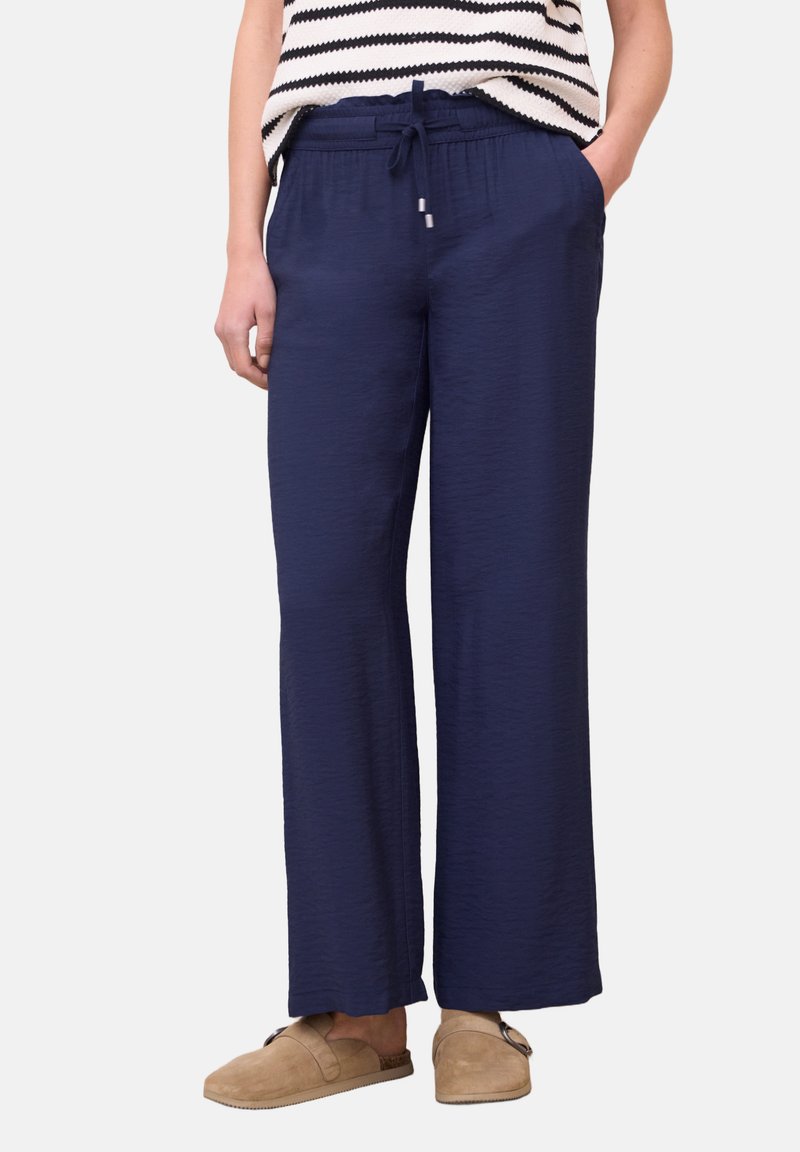 Femme portant un pantalon large bleu marine avec une taille élastique et un cordon, associé à des chaussures beige à enfiler et un haut rayé noir et blanc.