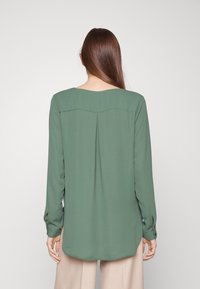 Chemise à manches longues en tissu vert doux, avec un col rond, des plis dans le dos et des poignets boutonnés.