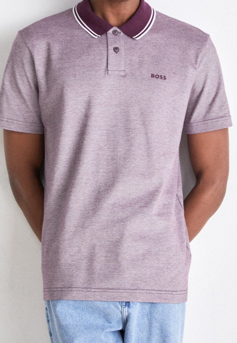 Polo en tissu violet clair avec un col violet foncé et des rayures blanches. Il possède trois boutons et un petit logo brodé.