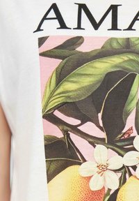 Hvid t-shirt med et grafisk design af grønne blade, gule frugter og hvide blomster på en pink baggrund. Teksten lyder "AMAZ."