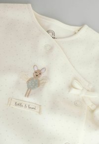 Cremefarbenes Kleidungsstück mit kleinen beigefarbenen Punkten, verziert mit einer gestickten Maus mit Flügeln. Enthält eine Schleifendetail und ein Etikett mit der Aufschrift "little & loved".