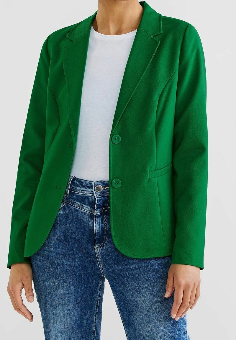 Zielony blezer o dopasowanym kroju, z dwoma guzikami i klapami. Noszony na białej koszulce, w połączeniu z niebieskimi dżinsami. Gładka faktura materiału.